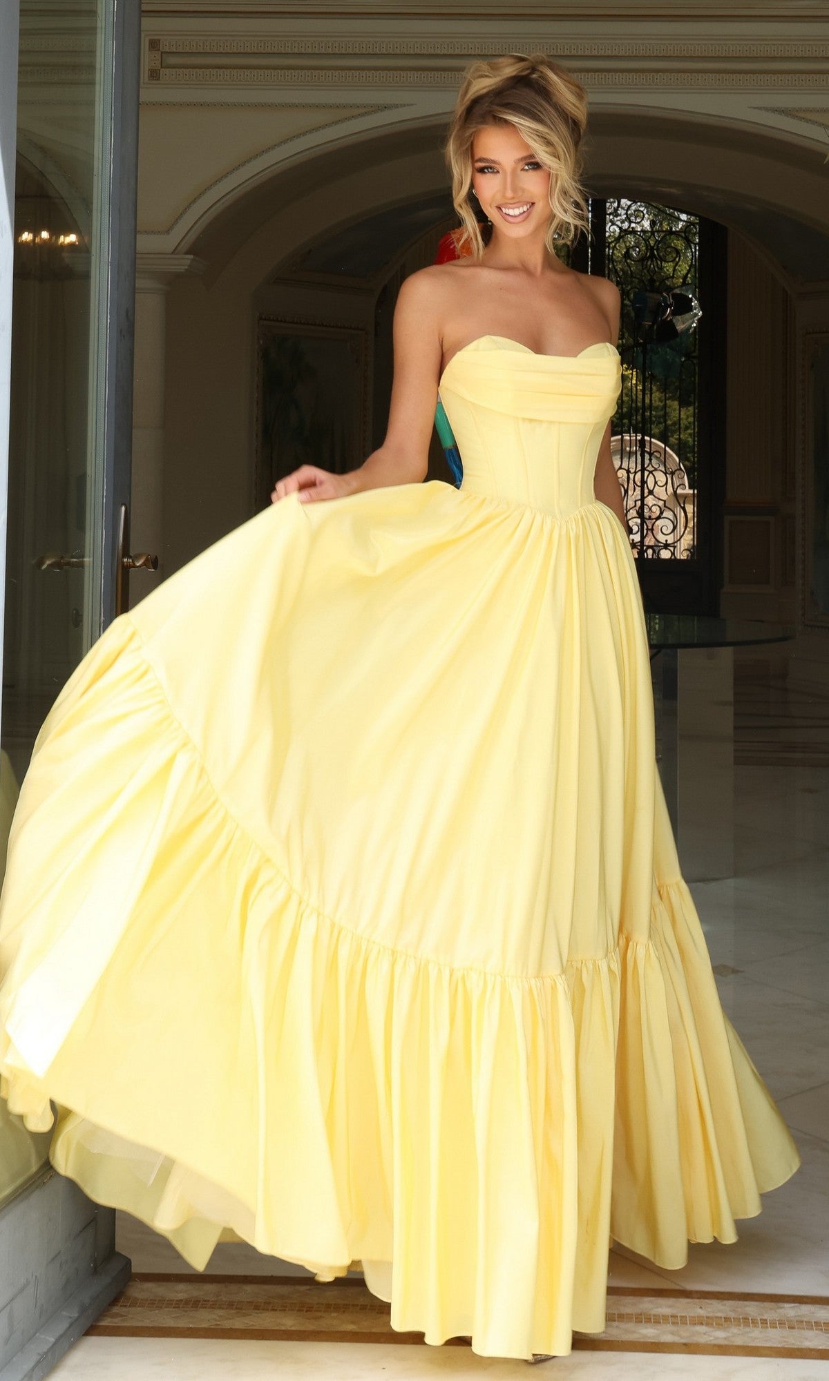 A woman in the Jovani 47883 Strapless Long Ruffled Prom Dress in yellow stands indoors, holding out one side of the skirt and smiling at the camera.